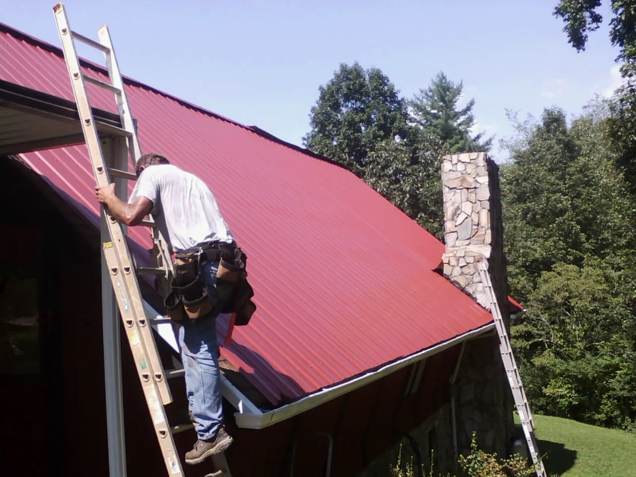 Roofer on a ladder beside a red metal roof and stone chimney — Soffit & Fascia Repair by Roofing Brothers in Angels Camp
